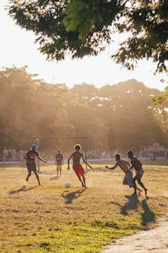 A group of young people playing soccer on a grassy field, with sunlight filtering through trees in the background. The scene captures the dynamic movement and energy of the game as dust is kicked up from the ground.