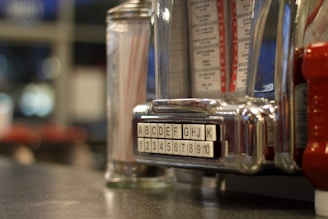 A close-up of a vintage jukebox selector. Alphabetic and numeric buttons are displayed prominently, with a reflective, chrome finish. In the background, blurred condiments like a glass container with straws and possibly a ketchup bottle are visible, evoking a retro diner atmosphere.