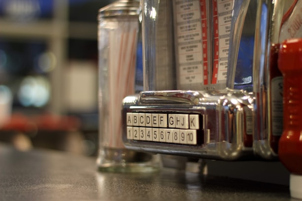 A close-up of a vintage jukebox selector. Alphabetic and numeric buttons are displayed prominently, with a reflective, chrome finish. In the background, blurred condiments like a glass container with straws and possibly a ketchup bottle are visible, evoking a retro diner atmosphere.