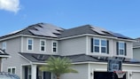 A modern two-story house with several solar panels installed on the roof. The exterior is light grey with white trims and black shutters. A small palm tree is visible in the front yard, and a basketball hoop is situated on the driveway. The sky is clear and blue, indicating fair weather.