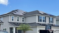 A modern two-story house with several solar panels installed on the roof. The exterior is light grey with white trims and black shutters. A small palm tree is visible in the front yard, and a basketball hoop is situated on the driveway. The sky is clear and blue, indicating fair weather.