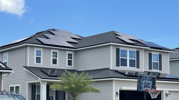 A modern two-story house with several solar panels installed on the roof. The exterior is light grey with white trims and black shutters. A small palm tree is visible in the front yard, and a basketball hoop is situated on the driveway. The sky is clear and blue, indicating fair weather.