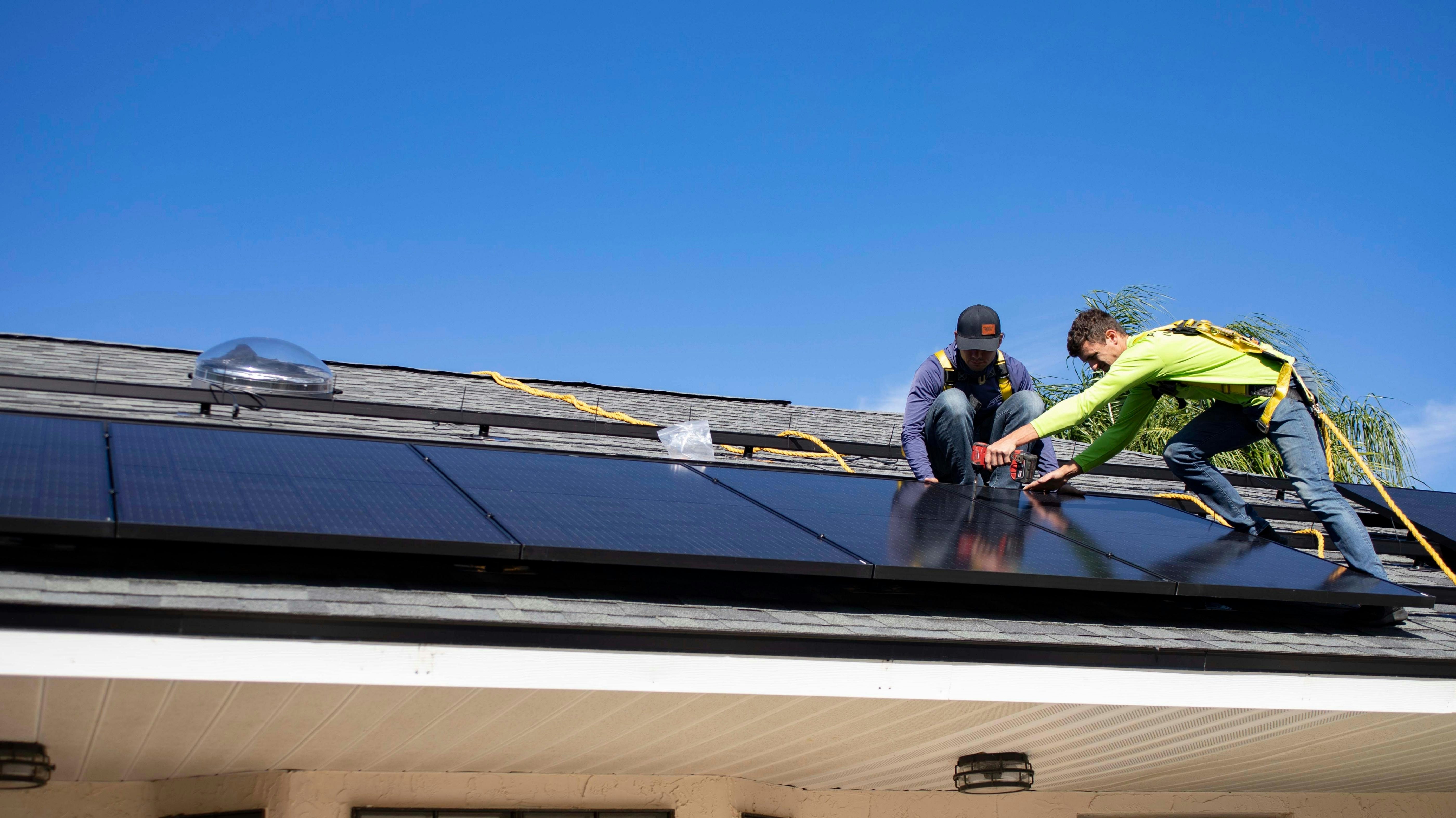 Two workers are installing solar panels on a rooftop under a clear blue sky. They are wearing safety harnesses and working with tools to secure the panels. The roof has a light gray shingle texture, and there is greenery in the background.