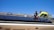 Two workers are installing solar panels on a rooftop under a clear blue sky. They are wearing safety harnesses and working with tools to secure the panels. The roof has a light gray shingle texture, and there is greenery in the background.