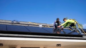 Two workers are installing solar panels on a rooftop under a clear blue sky. They are wearing safety harnesses and working with tools to secure the panels. The roof has a light gray shingle texture, and there is greenery in the background.