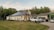 A modern house with solar panels on the roof is surrounded by a well-maintained lawn. Several vehicles are parked in the driveway, including a white truck and a car. The house is located near a wooded area with lush green trees in the background. The sky appears clear with a few clouds.