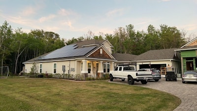 A modern house with solar panels on the roof is surrounded by a well-maintained lawn. Several vehicles are parked in the driveway, including a white truck and a car. The house is located near a wooded area with lush green trees in the background. The sky appears clear with a few clouds.