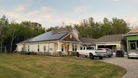 A modern house with solar panels on the roof is surrounded by a well-maintained lawn. Several vehicles are parked in the driveway, including a white truck and a car. The house is located near a wooded area with lush green trees in the background. The sky appears clear with a few clouds.