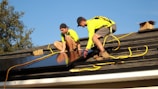 Two workers in bright yellow shirts are installing solar panels on a residential rooftop. They are using safety ropes and wearing protective gear. The scene is sunny with a clear blue sky, and a tree is visible in the background.