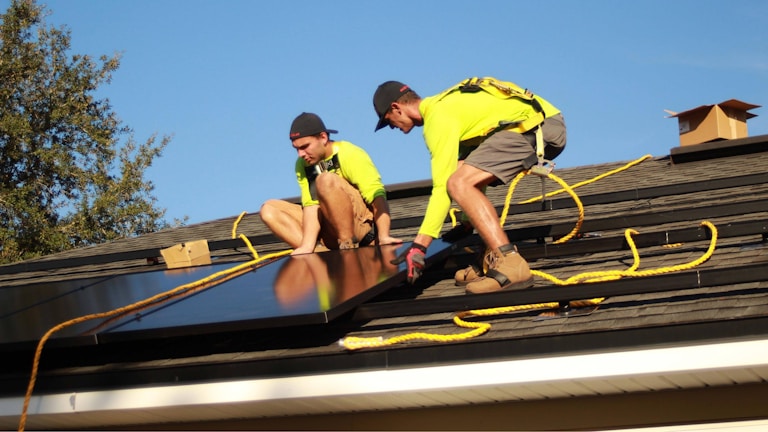 Two workers in bright yellow shirts are installing solar panels on a residential rooftop. They are using safety ropes and wearing protective gear. The scene is sunny with a clear blue sky, and a tree is visible in the background.