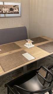 A dining setup featuring a wooden table with woven placemats, napkins, and a tissue holder with toothpicks. The surroundings include a cushioned bench and a framed monochrome cityscape photograph on the wall. The area exhibits a clean and organized setting.
