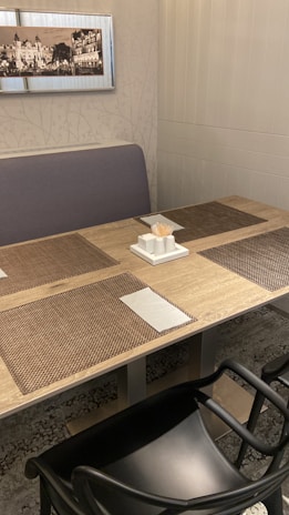 A dining setup featuring a wooden table with woven placemats, napkins, and a tissue holder with toothpicks. The surroundings include a cushioned bench and a framed monochrome cityscape photograph on the wall. The area exhibits a clean and organized setting.