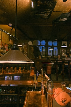 A cozy bar with dim lighting and wooden interiors. The bar counter is prominent with several beer taps and a glass of beer. A ceiling lamp hangs over the counter, adding to the warm ambiance. Patrons are visible in the background, seated and enjoying the atmosphere.