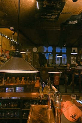 A cozy bar with dim lighting and wooden interiors. The bar counter is prominent with several beer taps and a glass of beer. A ceiling lamp hangs over the counter, adding to the warm ambiance. Patrons are visible in the background, seated and enjoying the atmosphere.