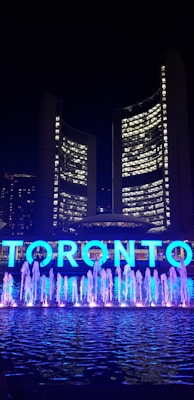 A nighttime scene featuring a large, illuminated sign spelling 'TORONTO' in front of a modern building. Bright blue lights highlight a row of water fountains in the foreground. The building in the background showcases numerous windows lit up, suggesting activity inside.