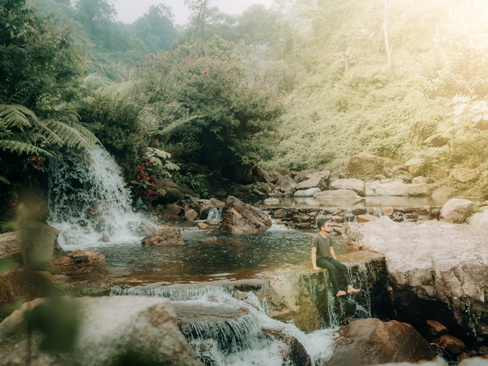 a person sitting on a rock by a river with a waterfall