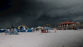 A beach scene with dark, ominous storm clouds looming overhead. The sandy foreground hosts several groups of people under blue and white striped umbrellas. On the left, there is a large inflatable water slide. To the right, a building with a red roof is visible, and palm trees are scattered in the background.