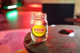 A clear glass jar labeled 'Smirnoff Lemonade' sits on a dark wooden surface. The background is blurred with vibrant, colorful lights in shades of green, red, and yellow, creating a lively atmosphere.