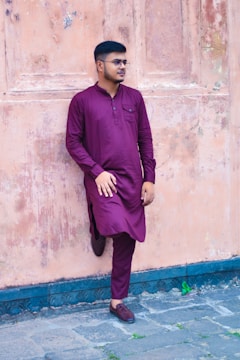 A person in a maroon traditional outfit stands casually against an aged, textured wall. The individual wears glasses and has one leg bent with a foot resting on the wall.