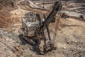 A large excavator is operating in a vast open-pit mining area, surrounded by dirt roads and rocky terrain. The machinery is actively digging into the earth, with dust rising from the operation. The landscape is vast and barren, showing multiple layers and pathways within the mine.