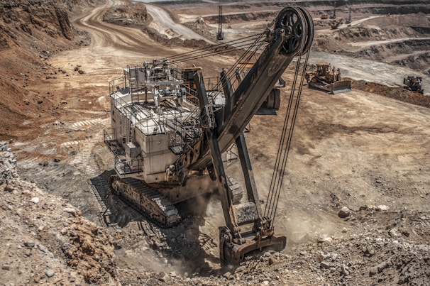 A large excavator is operating in a vast open-pit mining area, surrounded by dirt roads and rocky terrain. The machinery is actively digging into the earth, with dust rising from the operation. The landscape is vast and barren, showing multiple layers and pathways within the mine.