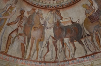 A mural depicting a historical scene with chariots and horses, along with people engaged in a procession or preparation. The art style and colors are reminiscent of ancient Roman or Greek frescoes.