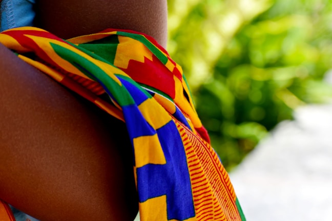 A vibrant, multicolored fabric, possibly traditional or cultural, draped over a person's arm. The background features a lush green, out-of-focus foliage.