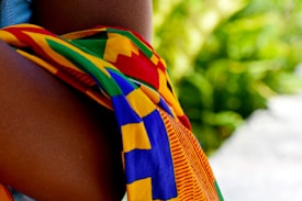 A vibrant, multicolored fabric, possibly traditional or cultural, draped over a person's arm. The background features a lush green, out-of-focus foliage.