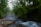 A lush, green forest scene with a flowing river and a natural hot spring pool. Mist hangs in the air, and two people wearing outdoor gear are walking along the riverbank.