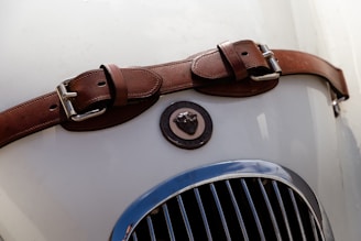 A brown leather strap with metal buckles is placed across the hood of a vintage car, featuring an emblem with a jaguar head. The grille of the car is visible and has vertical chrome slats.