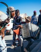 A group of people gathered outdoors, enjoying music played on an accordion. One person sits on a chair playing the accordion, while others stand around, some smiling and interacting. They are dressed casually, with a car and houses visible in the background under a clear blue sky.