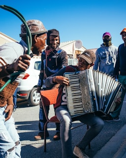 A group of people gathered outdoors, enjoying music played on an accordion. One person sits on a chair playing the accordion, while others stand around, some smiling and interacting. They are dressed casually, with a car and houses visible in the background under a clear blue sky.
