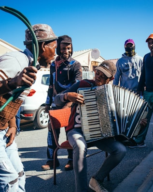 A group of people gathered outdoors, enjoying music played on an accordion. One person sits on a chair playing the accordion, while others stand around, some smiling and interacting. They are dressed casually, with a car and houses visible in the background under a clear blue sky.