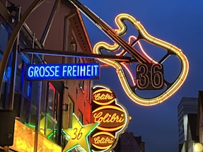 Neon signs illuminate a street scene with vibrant colors. A large neon guitar is prominently displayed with the number 36. Multiple signs are visible, including one with the words 'GROSSE FREIHEIT' in bright blue and another with 'Colibri' in red. The surrounding buildings are dimly lit, creating a lively nightlife atmosphere.