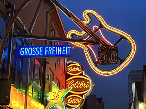 Neon signs illuminate a street scene with vibrant colors. A large neon guitar is prominently displayed with the number 36. Multiple signs are visible, including one with the words 'GROSSE FREIHEIT' in bright blue and another with 'Colibri' in red. The surrounding buildings are dimly lit, creating a lively nightlife atmosphere.