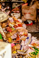 A person is engaged in a colorful craft activity, surrounded by vibrant paper fans or decorations. The hands are actively working with the materials, suggesting careful craftsmanship. The background contains various items, adding a sense of a busy, possibly festive environment.