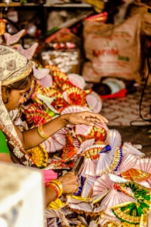 A person is engaged in a colorful craft activity, surrounded by vibrant paper fans or decorations. The hands are actively working with the materials, suggesting careful craftsmanship. The background contains various items, adding a sense of a busy, possibly festive environment.