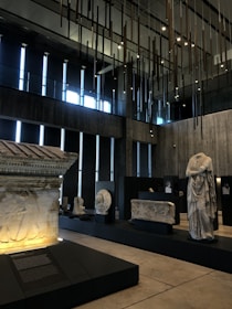 A modern interior space that features various ancient stone artifacts displayed on black platforms, with a detailed frieze and a headless statue among the pieces. Large vertical windows provide natural light, while a unique installation of hanging vertical rods decorates the ceiling. The overall ambiance is serene and contemplative.