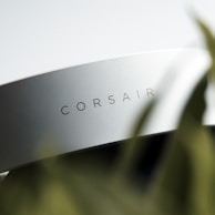 A close-up of a sleek, white object with the word 'CORSAIR' printed on its surface. The focus on the brand name suggests it is a piece of technology or an accessory. There is a blurred foreground element that appears to be a green plant, adding a natural contrast to the modern object.