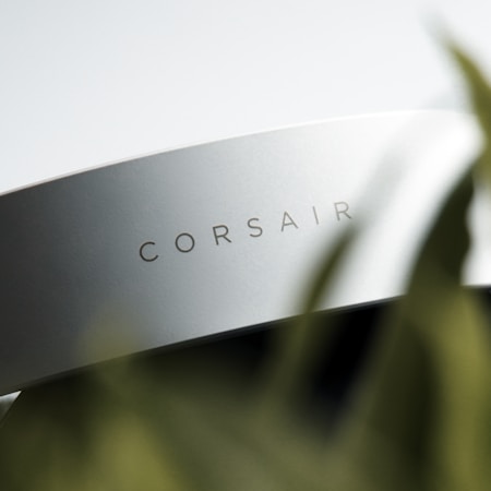 A close-up of a sleek, white object with the word 'CORSAIR' printed on its surface. The focus on the brand name suggests it is a piece of technology or an accessory. There is a blurred foreground element that appears to be a green plant, adding a natural contrast to the modern object.