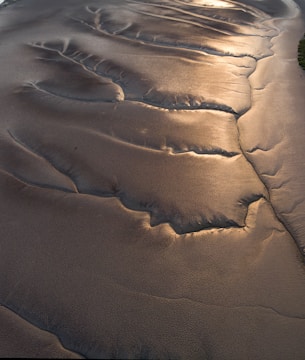 Rippling patterns in the sand are illuminated by soft, golden light, creating a textured and almost metallic appearance. The smooth surface is interrupted by natural formations, likely caused by water flow.