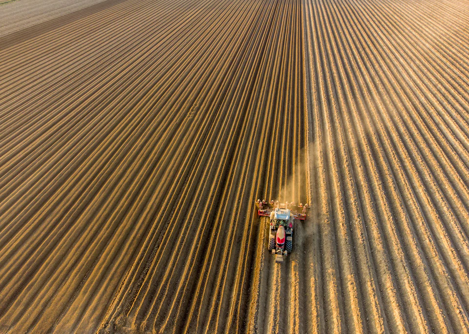 Large tractor plows central field from aerial perspective, leaving behind trail of disturbed earth
