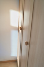 A wooden wardrobe with simple round knobs on the doors, bathed in soft natural light creating warm tones. The light casts a square shadow on the adjacent white wall, suggesting a nearby window.