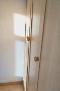 A wooden wardrobe with simple round knobs on the doors, bathed in soft natural light creating warm tones. The light casts a square shadow on the adjacent white wall, suggesting a nearby window.