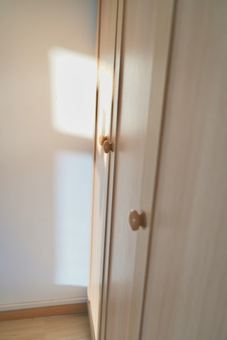 A wooden wardrobe with simple round knobs on the doors, bathed in soft natural light creating warm tones. The light casts a square shadow on the adjacent white wall, suggesting a nearby window.