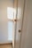 A wooden wardrobe with simple round knobs on the doors, bathed in soft natural light creating warm tones. The light casts a square shadow on the adjacent white wall, suggesting a nearby window.