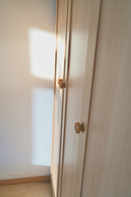 A wooden wardrobe with simple round knobs on the doors, bathed in soft natural light creating warm tones. The light casts a square shadow on the adjacent white wall, suggesting a nearby window.