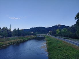A serene landscape with a flowing river bordered by green grass and trees. A road runs parallel to the river on the right side. In the background, a forested hill rises with a prominent building visible at the top. A church steeple with a green roof is seen on the left, set against an expansive blue sky.