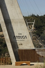 A large concrete structure labeled '516 Arouca Ponte Suspensa' appears prominently. The structure is part of a suspension bridge, with steel cables extending into the background. Wooden railings and a bench are at the base of the structure, and green hills are visible in the distance.