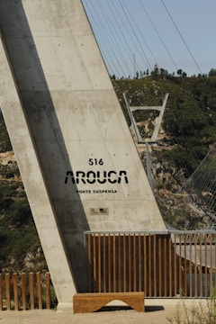A large concrete structure labeled '516 Arouca Ponte Suspensa' appears prominently. The structure is part of a suspension bridge, with steel cables extending into the background. Wooden railings and a bench are at the base of the structure, and green hills are visible in the distance.